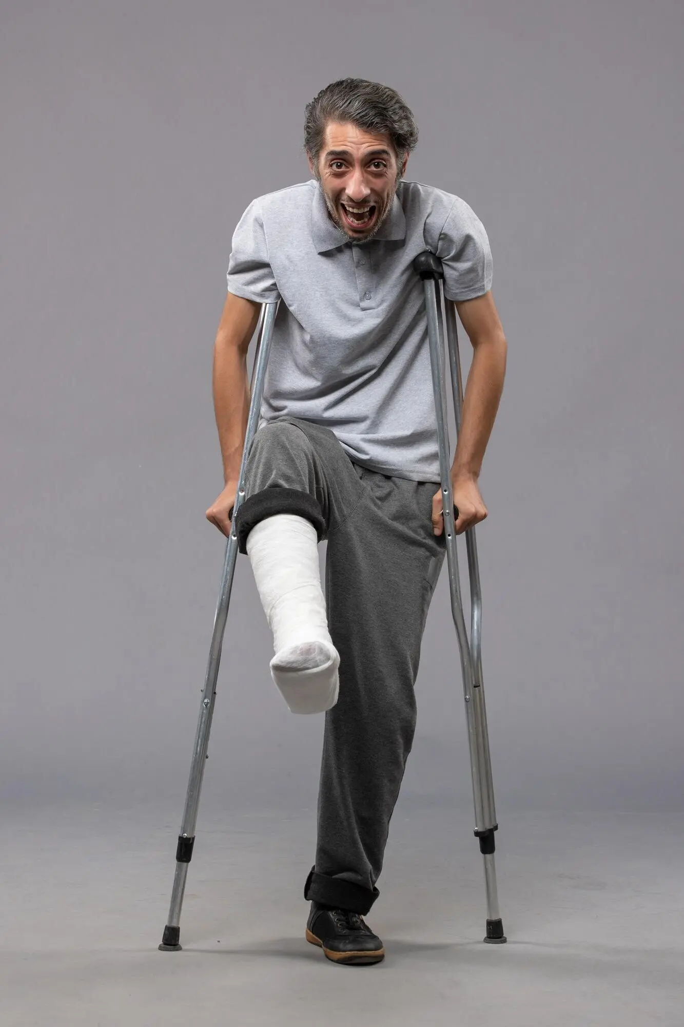 Front view of a young man using crutches because of a broken foot, on a grey desk; accident, disabled, broken leg.