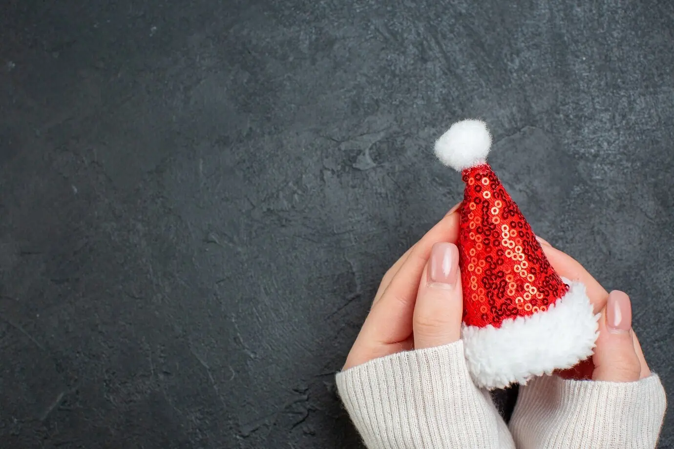 Overhead view of a hand holding a Santa Claus hat on the left side against a dark background.